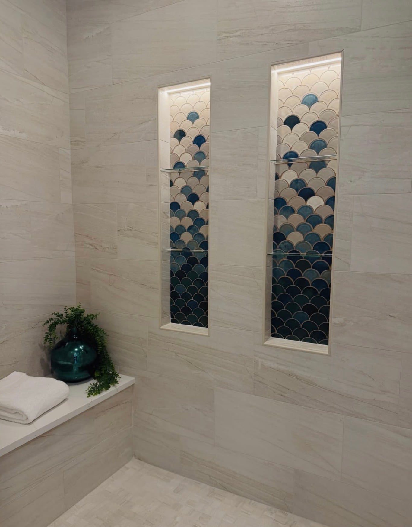 Luxury walk-in tile shower with two tall LED-lighted shower niches featuring blue and white ombré fish scale mosaic tiles, large beige stone-look wall tiles, glass shelves, and built-in shower bench.