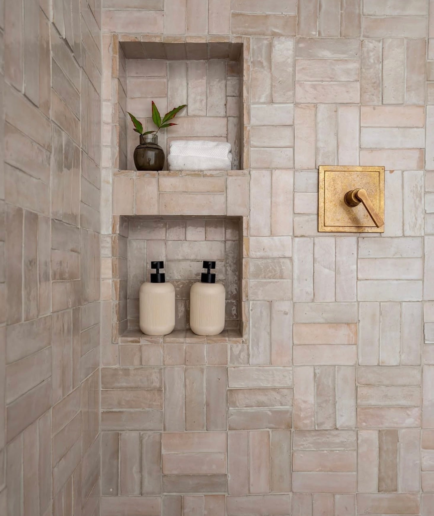 Rustic tile shower with beige and blush-toned rectangular tiles arranged in a basketweave pattern, two stacked recessed shower niches, textured handmade-look tile, and antique brass shower control fixture.