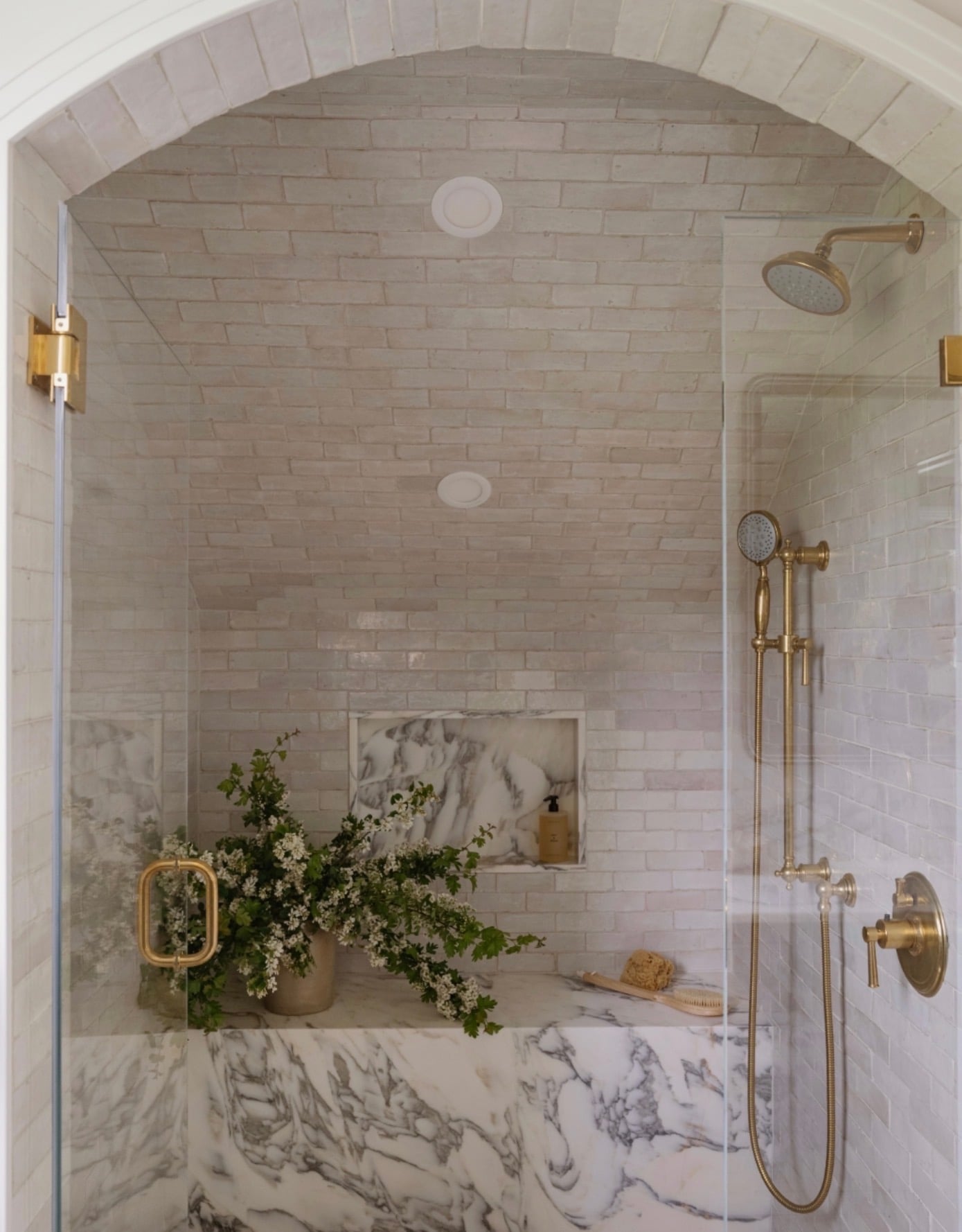 Luxury arched walk-in shower with white subway tile walls and vaulted ceiling, dramatic white and gray marble shower bench, matching marble shower niche, brushed gold rainfall showerhead, and frameless glass doors with gold hardware.
